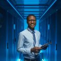 Waist up portrait of smiling African American man standing by server cabinet while working with supercomputer in data center and holding tablet, copy space